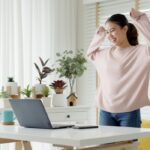 A female writer in her study wearing a pink jumper, looking at her laptop while standing up and cheering in celebration.